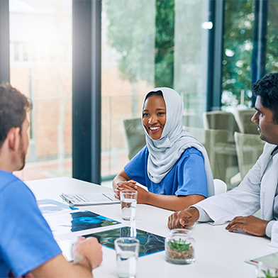 A group of culturally diverse nurses are seated at a conference table attending a meeting. They are smiling and engaged in conversation. A group of culturally diverse nurses are seated at a conference table attending a meeting. They are smiling and engaged in conversation.