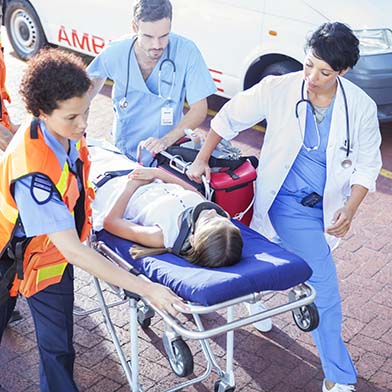 Two Emergency Room staff members are helping a team of emergency medical technicians to push a stretcher into the Emergency Department. A young child wearing a neck brace is calmly laying on the stretcher. Two Emergency Room staff members are helping a team of emergency medical technicians to push a stretcher into the Emergency Department. A young child wearing a neck brace is calmly laying on the stretcher.