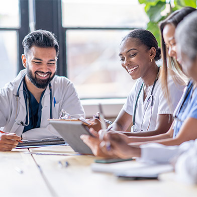 A small group of four medical professionals sit around a boardroom table as they meet to discuss patient cases. They are each dressed professionally in scrubs and lab coats as they focus on working together. A small group of four medical professionals sit around a boardroom table as they meet to discuss patient cases. They are each dressed professionally in scrubs and lab coats as they focus on working together.