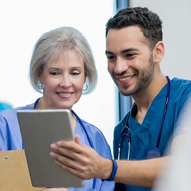 An older female nurse and a younger male nurse are standing together. The older nurse is reviewing some documentation that the younger nurse is holding in his hand and showing her. Both are smiling. An older female nurse and a younger male nurse are standing together. The older nurse is reviewing some documentation that the younger nurse is holding in his hand and showing her. Both are smiling.