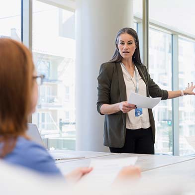 A female Director of Nursing stands in front of a conference table and is looking at a group of seated employees while giving a presentation and pointing to some information on a whiteboard behind her. A female Director of Nursing stands in front of a conference table and is looking at a group of seated employees while giving a presentation and pointing to some information on a whiteboard behind her.