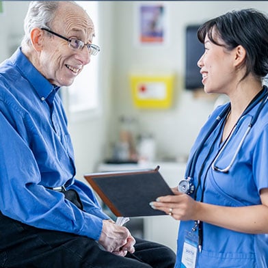 A nurse in a blue scrub top and badge provides coaching to a smiling elderly man in a blue shirt, holding a tablet in a clinic office, demonstrating a patient-centered approach in nursing. A nurse in a blue scrub top and badge provides coaching to a smiling elderly man in a blue shirt, holding a tablet in a clinic office, demonstrating a patient-centered approach in nursing.